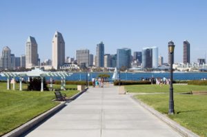 Image of San Diego skyline on a bright sunny afternoon. The sky is blue without any clouds. The grass is green and at the end of the parkway is a lake.